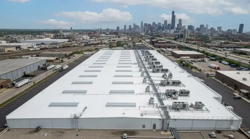 Aerial view of a large white membrane roof on a warehouse in an industrial corridor, skylights and rooftop units in rows, city skyline behind