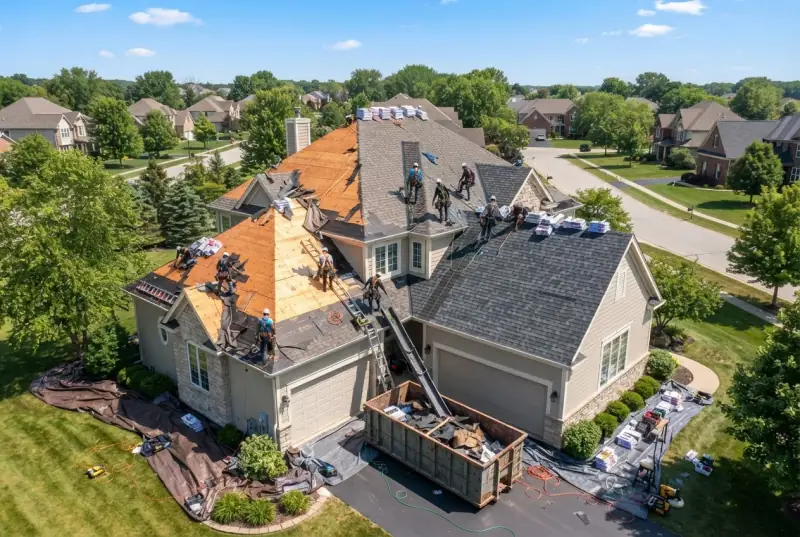 Aerial view of a large suburban house being reroofed, part of the slope down to bare decking, shingle bundles staged across the roof and a loaded skip on the drive